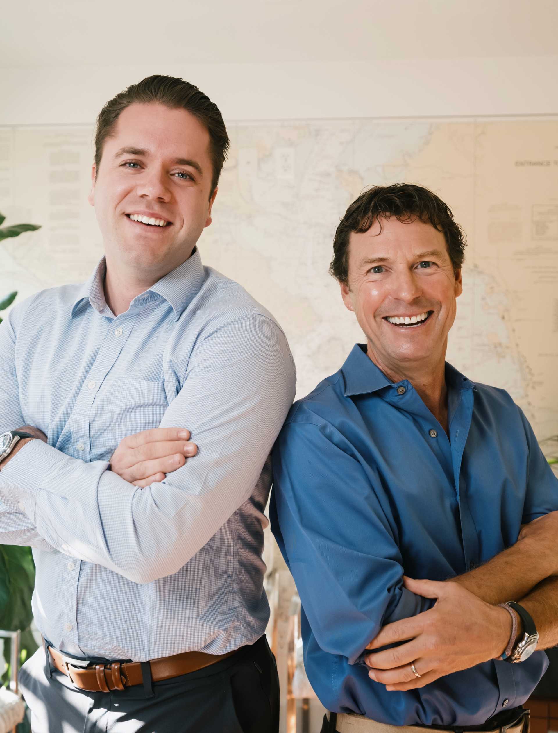 Two smiling men standing back to back with arms crossed, one in a light blue shirt and the other in a dark blue shirt.
